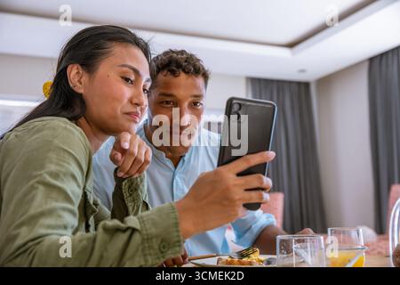 Couple diversifié penché à la table du petit déjeuner dans le coin salle à manger avec smartphone au-dessus de verres de jus Banque D'Images