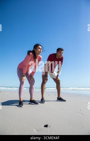Couple diversifié penché en avant reprenant son souffle sur le bord de mer dans des vêtements de sport et des chaussures de course Banque D'Images