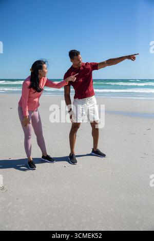 Couple diversifié penché en avant et pointant vers l'horizon de l'océan sur la plage de sable avec des chaussures Banque D'Images