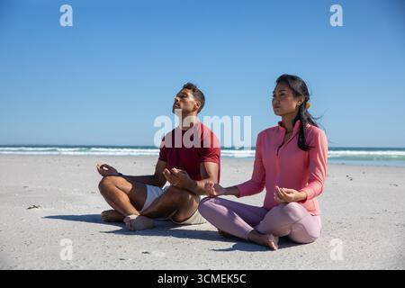 Couple diversifié méditant les jambes croisées sur la plage de sable en tenue athlétique sous le ciel bleu Banque D'Images