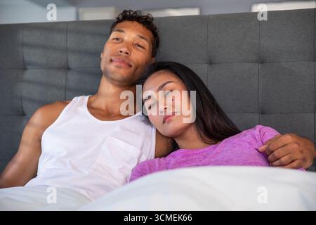 Couple diversifié incliné sous couette contre tête de lit rembourrée grise sur le lit dans la chambre Banque D'Images