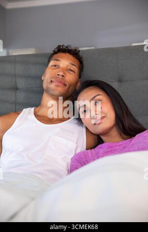 Couple diversifié s'inclinant sur un grand lit avec des oreillers blancs contre tête de lit grise dans la chambre Banque D'Images