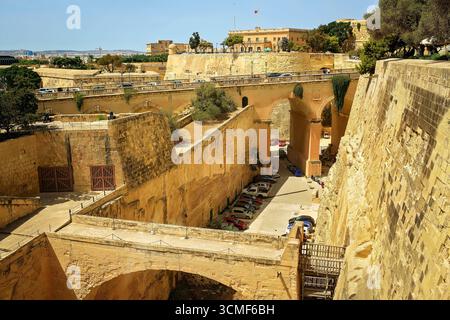 Vue panoramique sur les anciennes fortifications en pierre et les ponts voûtés à la Valette, Malte, avec des voitures garées près des murs de la forteresse et vue sur le paysage urbain. Banque D'Images