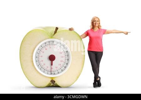 Portrait pleine longueur d'une femme mature en vêtements de sport posant à côté d'une échelle de mesure de pomme et pointant isolé sur fond blanc Banque D'Images