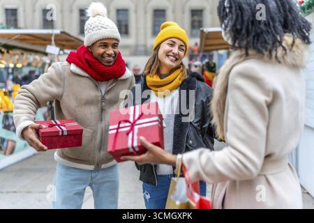 Heureux amis échangeant des cadeaux de noël sur un marché animé, profitant de l'atmosphère festive et célébrant la joie de la saison Banque D'Images