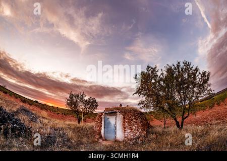 Une vue légèrement surélevée et grand angle d'une petite cabane ronde en pierre avec une porte altérée. La cabane se dresse dans un champ d'herbes sèches et sauvages et de soi rougeâtre Banque D'Images