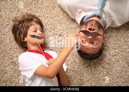 Père et fils émotionnels avec moustache en papier couché sur un tapis, vue de dessus Banque D'Images
