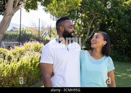Couple diversifié debout dans le parc ensoleillé serrant à la taille par la clôture de maillons de chaîne près des arbustes à fleurs Banque D'Images
