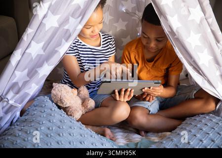 Divers frères et sœurs d'enfant explorant la tablette à l'intérieur du tipi étoilé avec ours en peluche dans le salon Banque D'Images