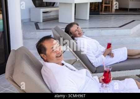 Couple asiatique sénior assis sur des chaises de patio portant des peignoirs en sirotant une boisson dans les verres Banque D'Images