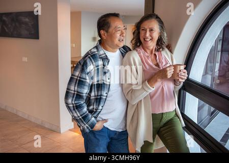 Couple senior diversifié debout par la fenêtre à la maison tenant une tasse en céramique et regardant à l'extérieur Banque D'Images