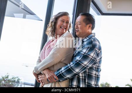 Couple senior diversifié debout près des portes vitrées serrant sur le balcon de bord de mer sous le ciel ensoleillé Banque D'Images