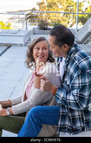 Couple diversifié tenant une tasse à café et échangeant des sourires sur la terrasse avec des plantes en pot Banque D'Images