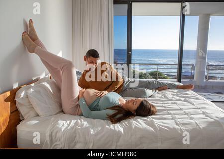Couple diversifié allongé sur le lit soutenant la bosse de bébé à côté des portes coulissantes en verre donnant sur l'océan calme Banque D'Images