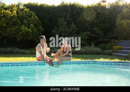 Couple assis au bord de la piscine dans la cour arrière trempant les pieds dans l'eau carrelée tout en tenant des tasses Banque D'Images