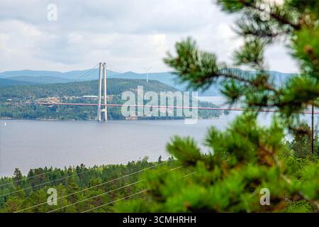 High Coast Bridge, une structure à haubans de câbles en Suède, enjambe une étendue d'eau, reliant des rivages sous un ciel nuageux, avec des éoliennes lointaines et f Banque D'Images