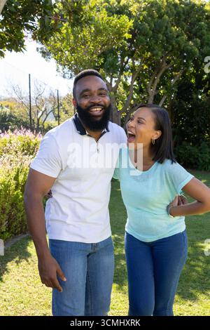 Couple diversifié debout dans le jardin ensoleillé sur la pelouse herbeuse penché l'un vers l'autre riant Banque D'Images