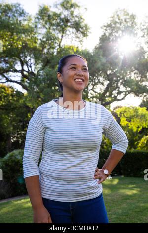 Femme asiatique souriante et regardant vers le haut dans le jardin ensoleillé portant chemise rayée, jeans et montre-bracelet Banque D'Images