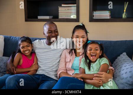 Famille diversifiée assis sur le canapé bleu à la maison souriant par des étagères avec des livres et bouteille de diffuseur Banque D'Images