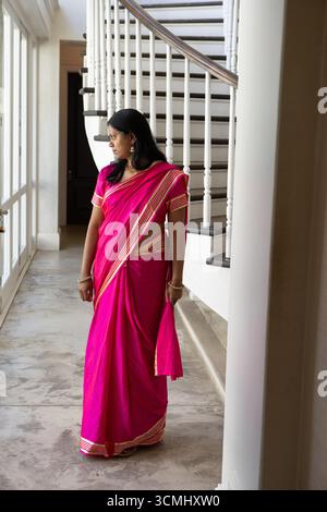 Femme indienne debout dans le foyer, portant saree rose vif et bijoux en or par de grandes fenêtres Banque D'Images