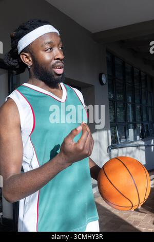 Homme afro-américain debout sous une passerelle couverte, portant un maillot vert tenant le basket-ball Banque D'Images