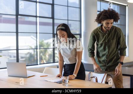 Divers collègues debout à la table de conférence examinant des documents imprimés à l'aide d'un ordinateur portable Banque D'Images