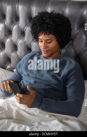 Homme afro-américain utilisant un smartphone assis sur le lit, tête de lit en cuir touffeté, literie blanche Banque D'Images