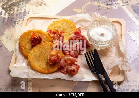 Une portion de crêpes de pommes de terre dorées, frites avec bacon croustillant et sauce, servies sur une planche de bois avec du papier artisanal et des couverts. Banque D'Images