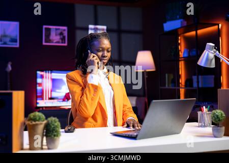 Télétravailleur afro-américain recevant un appel téléphonique d'un collègue de travail tout en travaillant à domicile sur un ordinateur portable. Freelance parlant avec un collègue à l'aide d'un smartphone tout en résolvant des tâches Banque D'Images
