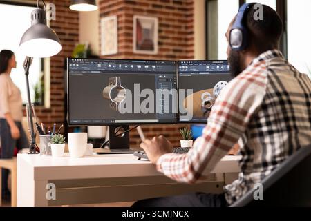 Homme afro-américain avec des écouteurs, analysant la conception sur le moniteur à l'aide d'un logiciel de cao. Concepteur à distance innovant écoutant de la musique et travaillant sur la simulation d'outils de construction depuis la maison de mur de briques. Banque D'Images