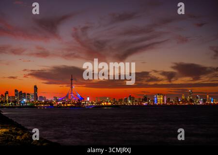 Vue imprenable au crépuscule sur les gratte-ciel illuminés de Koweït City, avec la Marina Crescent Sailing Tower qui brille contre un coucher de soleil vibrant sur l'Ara Banque D'Images