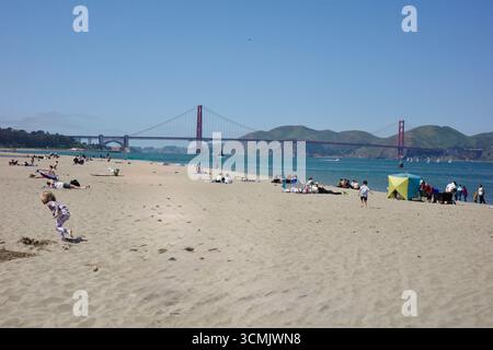 Les gens se détendent sur Crissy Field Beach à San Francisco, avec des enfants jouant dans le sable et le Golden Gate Bridge s'élevant en arrière-plan sous un b clair Banque D'Images