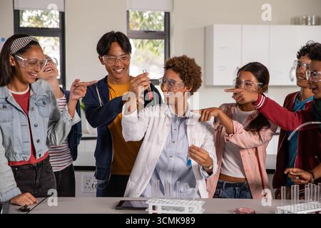 Femme afro-américaine en blouse de laboratoire tenant un tube à essai de solution rouge guidant des étudiants adolescents au banc Banque D'Images