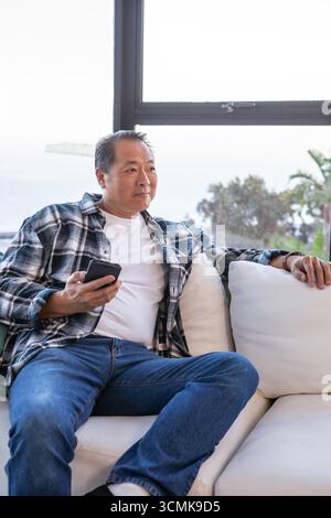 Homme chinois asiatique senior assis sur le canapé dans le salon tenant smartphone Banque D'Images