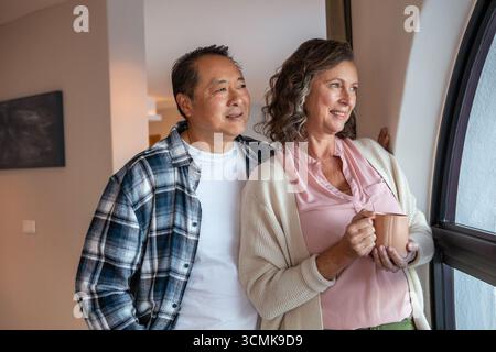 Couple senior debout derrière partenaire regardant par la fenêtre cintrée tenant une tasse en céramique à la maison Banque D'Images