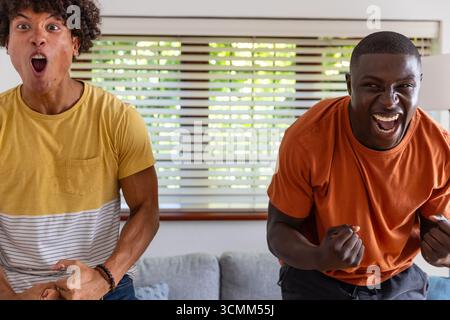 Divers amis masculins acclamant et se penchant en avant dans le salon près du canapé avec des oreillers décoratifs Banque D'Images