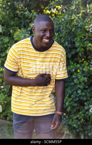 Homme afro-américain debout dans le jardin portant un tee-shirt rayé montrant montre-bracelet et riant Banque D'Images