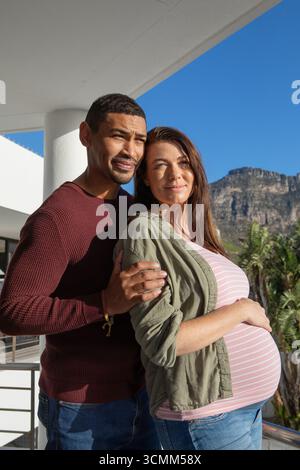 Couple enceinte adulte moyen embrassant sur le balcon appuyé sur la balustrade surplombant les montagnes et les palmiers Banque D'Images