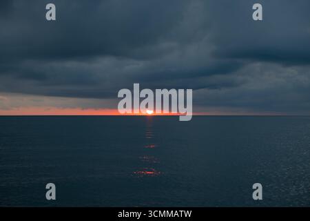 Lever de soleil rouge se couchant sur la mer sous de lourds nuages sombres. Scène de coucher de soleil Moody et intense avec une ligne d'horizon droite sur la mer calme Banque D'Images