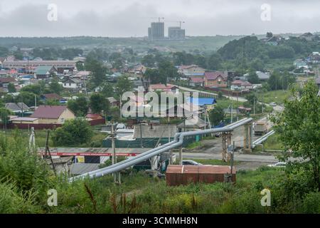 Pipelines surélevés et panorama sur les toits résidentiels à Korsakov, dans la région russe de Sakhaline, avec grues de construction sur une pente verdoyante. Banque D'Images