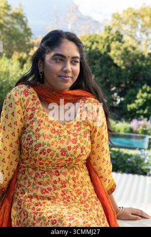 Femme indienne assise sur le banc dans la terrasse du jardin, portant kurta jaune avec dupatta orange près de la piscine Banque D'Images