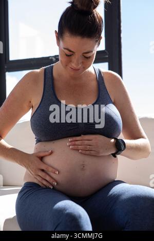 Femme adulte moyenne assise sur un canapé léger près de la fenêtre, berçant le ventre enceinte et montrant smartwatch Banque D'Images