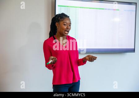 Femme afro-américaine en blouse présentant feuille de calcul sur écran dans le bureau tenant marqueur Banque D'Images