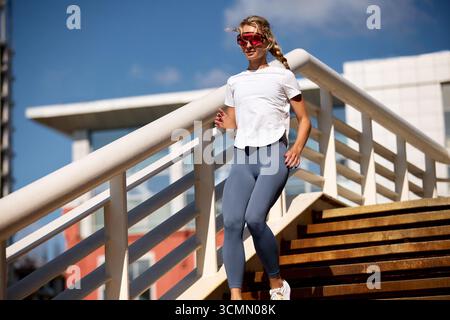 Une jeune femme aime une séance d'entraînement matinale alors qu'elle court à l'extérieur sur un escalier portant des vêtements de sport à la mode, l'air concentrée et énergique. Son activité est élevée Banque D'Images