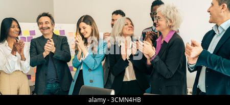 Une équipe heureuse et diversifiée de professionnels des affaires multigénérationnels se tient et applaudit ensemble, célébrant une réussite lors d'un corpor Banque D'Images