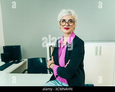 Une femme d'affaires senior confiante et prospère avec des lunettes élégantes et des cheveux gris courts sourires tout en posant avec ses bras croisés, assise sur ses des Banque D'Images