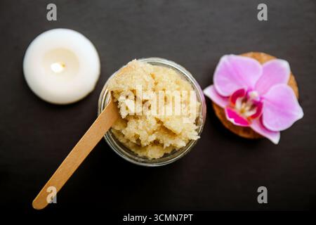 Au-dessus de la vue du gommage du corps en sucre fait maison dans un pot en verre, décoré avec fleur d'orchidée de couleur rose et bougie de spa brûlante, fond de pierre noire. Banque D'Images