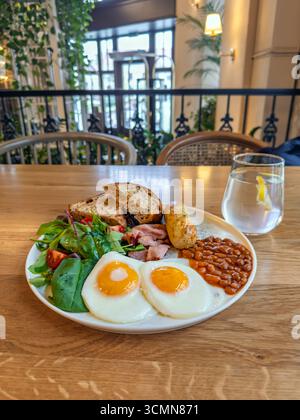 Petit déjeuner anglais traditionnel avec œufs au plat, bacon, haricots, saucisses et salade servis sur une assiette blanche dans un restaurant confortable. Petit déjeuner de style britannique. Banque D'Images