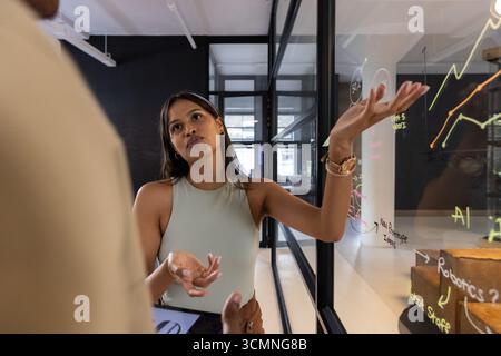 Divers collègues faisant des gestes sur des cartes murales en verre coloré, discutant des données dans le couloir du bureau Banque D'Images