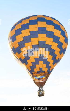 Ballon à air chaud flottant dans le ciel dégagé avec un adulte caucasien debout dans les commandes de fonctionnement du panier, panier suspendu sous une grande enveloppe de ballon à damier jaune et bleu Banque D'Images